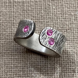 May include: An open-style silver ring featuring a textured, dark oxidized finish on the outer surface. The ring is accented with three small, round, vibrant pink gemstones set into the metal, with one on one side and two on the other.