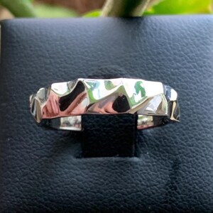 Könnte beinhalten: Ein silberfarbener Ring mit einem strukturierten, geometrischen Design. Der Ring hat eine Reihe von abgewinkelten Facetten, die das Licht reflektieren und einen schimmernden Effekt erzeugen. Der Ring wird auf einer schwarzen Oberfläche präsentiert.