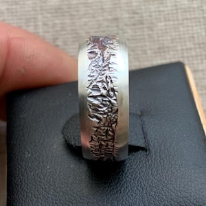 Peut inclure: Une bague argentée dotée d'une large bande avec des bords lisses et brossés, ainsi qu'un motif central en relief, sombre, profondément texturé et accidenté, qui s'étend sur toute la circonférence de l'anneau, créant un contraste marqué.