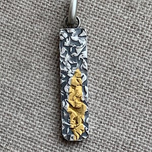 May include: A rectangular pendant featuring a dark, oxidized silver surface with a raised, textured floral pattern. A vertical, irregular strip of bright yellow gold is centered on the pendant. The piece includes a silver bail at the top for hanging.