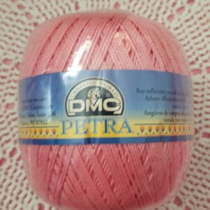 May include: A ball of pink DMC Petra crochet thread, wrapped in clear plastic. The label features the DMC logo and the word "PETRA" in yellow. The thread is displayed on a white and pink crocheted doily.
