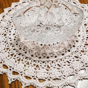 May include: A clear, cut-glass basket with a handle sits atop a white, crocheted doily. The basket has a detailed, geometric pattern. The doily is round with a scalloped edge, resting on a wooden surface.