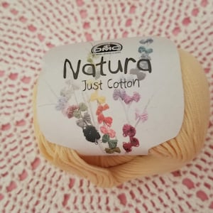 May include: A ball of Natura Just Cotton yarn in a soft yellow hue, with a label featuring the brand name and floral illustrations. The yarn is set against a pink and white crocheted background, creating a contrast of textures.