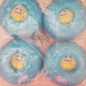 May include: Four balls of DMC Petra size 3 yarn in a light blue color, packaged in clear plastic. Each ball has a yellow label with the DMC Petra logo and weight information. The yarn is displayed on a white lace doily.