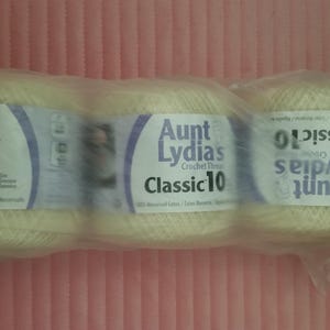May include: A three-pack of Aunt Lydia's Classic 10 crochet thread in a cream color, wrapped in clear plastic packaging. Each spool features a white label with purple text and graphics, clearly displaying the brand name and product information.