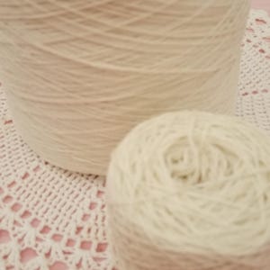 May include: A large cylindrical spool of off-white yarn positioned behind a smaller, round ball of the same off-white yarn. Both items feature a consistent, fine-textured fiber construction in a neutral, light cream color throughout the entire surface.