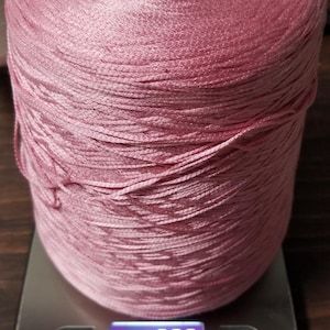 May include: A large spool of shiny, light pink thread or yarn resting on a digital kitchen scale. The scale display shows a weight of 750 grams. The spool has a cylindrical shape with a hollow cardboard center and a smooth, reflective texture.