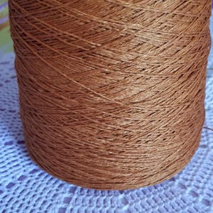 May include: A large cylindrical spool of metallic copper-colored yarn featuring a smooth, shiny texture and a tightly wound cross-hatch pattern. The thread exhibits a warm, reflective finish throughout the entire surface of the spool.