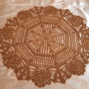 May include: A round, light brown crochet doily featuring an intricate, openwork geometric pattern. The design radiates from a central point, expanding into a series of interconnected loops and stitches that form a detailed, scalloped edge.