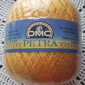 May include: A golden-yellow ball of DMC Petra crochet thread. The label reads "CREATIVE WORLD DMC PETRA" with additional text. The thread is tightly wound and packaged in clear plastic, suitable for various crafting projects.
