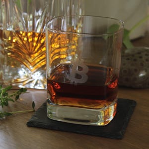 May include: A rocks glass with a Bitcoin symbol etched on the side, filled with amber liquid, sits on a black square coaster. A cut-glass decanter and sprigs of greenery are in the background. The glass is on a wooden surface.