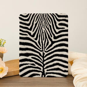 Puede incluir: Una funda rectangular para tableta con un patrón de rayas de cebra en blanco y negro de alto contraste. El diseño consiste en líneas orgánicas y fluidas que convergen hacia el centro, cubriendo toda la superficie frontal de la funda.