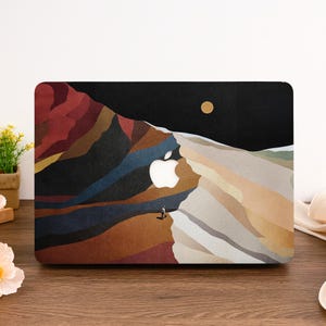 May include: Laptop case featuring a mountain landscape design with a small figure and a golden sun against a black sky. The Apple logo is integrated into the mountain design. The color palette includes shades of red, brown, and beige.