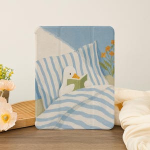 May include: Tablet case with a charming illustration of a duck reading in bed. The design features blue and white striped bedding and a light blue backdrop. The case is displayed on a wooden surface.