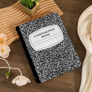Peut inclure: Un cahier de composition avec une couverture à motifs mouchetés noirs et blancs. La couverture comporte une étiquette blanche avec les mots "COMPOSITION BOOK" imprimés en noir. Le cahier est posé sur une surface en bois.