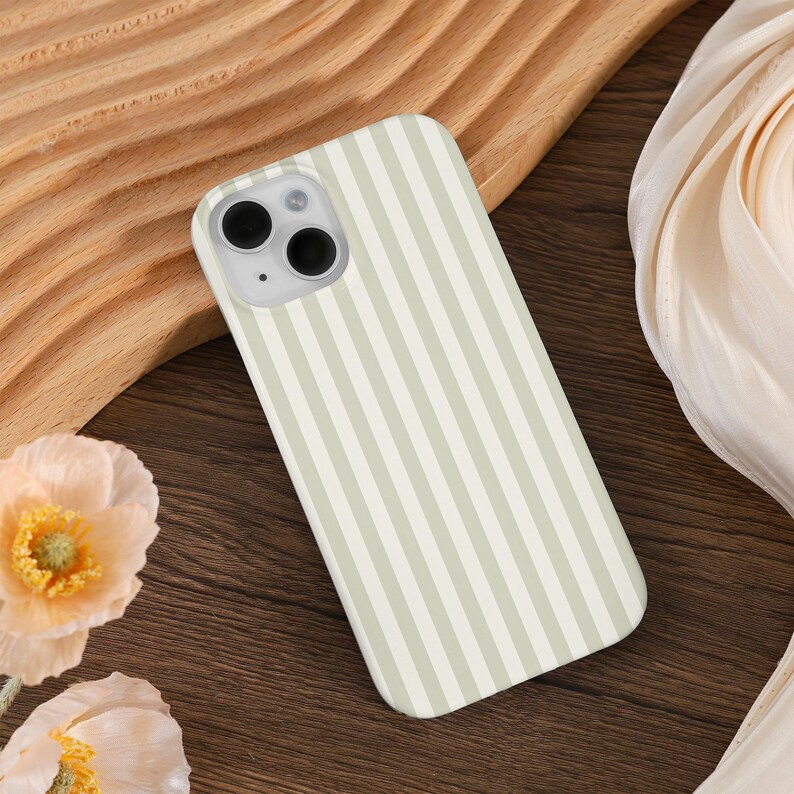 May include: A smartphone case with a vertical striped pattern in light green and white. The case has a camera cutout and is resting on a wooden surface. The overall aesthetic is minimalist and modern.