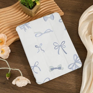 Könnte beinhalten: Weiße iPad-Hülle mit einem Muster aus blauen und weißen Schleifen. Die Hülle hat einen weißen Hintergrund mit blauen Schleifen in verschiedenen Größen und Formen.