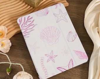 Può includere: Una custodia bianca per iPad con un design a conchiglie e stelle marine rosa e bianche. La custodia ha uno sfondo bianco con contorni rosa di conchiglie, stelle marine e alghe.