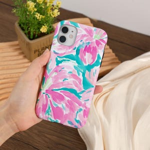 May include: A floral phone case featuring a vibrant watercolor design in shades of pink, teal, and white. The case has a glossy finish and a black border. The design showcases large, abstract flower shapes.