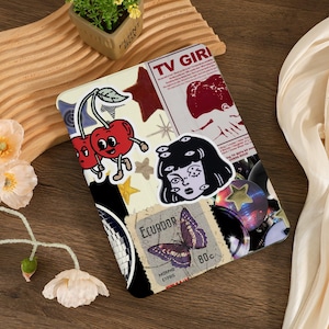 May include: A tablet case with a collage design featuring cartoon cherries, a woman with multiple eyes, stars, and a butterfly stamp from Ecuador. The text "TV GIRL" is visible on the case.