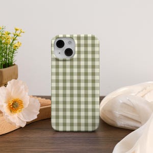 May include: A phone case featuring a green and white gingham design. The case has a camera aperture and is placed on a wooden surface. A white flower and a small plant in a wooden pot are in the background.