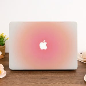 Könnte beinhalten: Eine Laptop-Hülle mit einem Farbverlauf in Rosa und Pfirsich, in der Mitte mit einem weißen Apple-Logo. Die Hülle hat eine glatte Oberfläche und ist für einen Laptop konzipiert.
