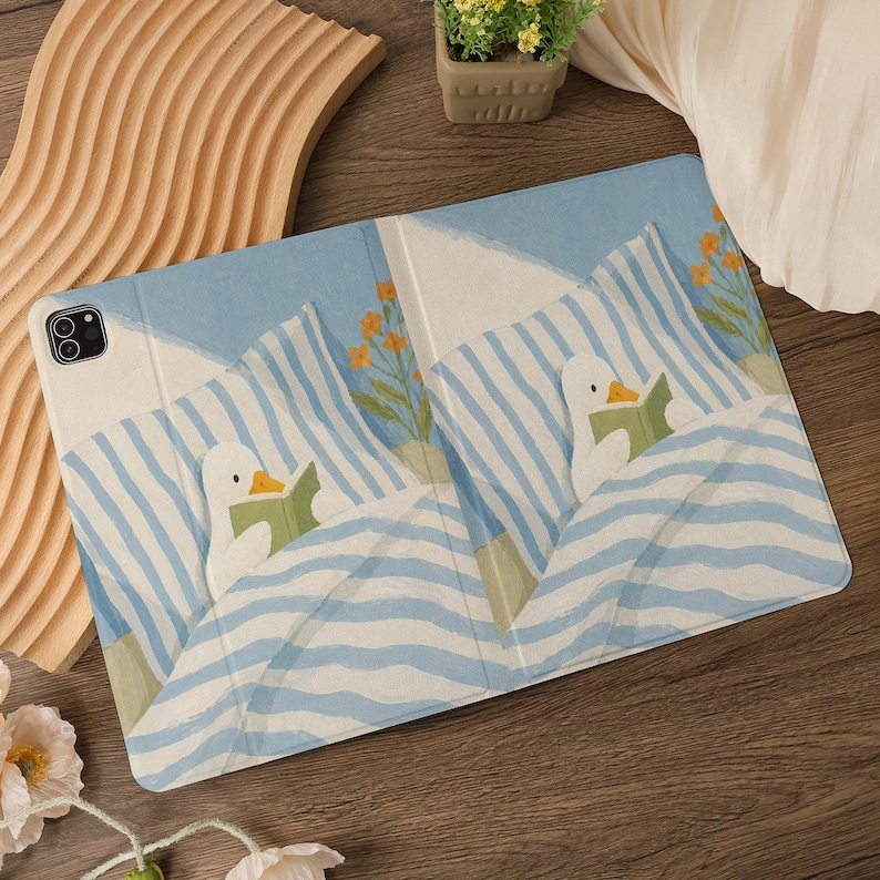 Puede incluir: Funda para tableta con un dise&ntilde;o de pato de dibujos animados leyendo en la cama, sobre un fondo de rayas azules y blancas. El dise&ntilde;o incluye flores naranjas. La funda es de color azul claro.