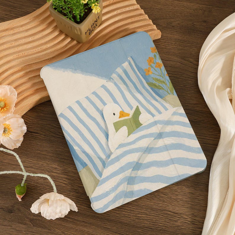 Puede incluir: Una funda para tableta con una ilustraci&oacute;n caprichosa de un pato leyendo en la cama. La funda presenta un patr&oacute;n de rayas azules y blancas, con un fondo azul claro y una peque&ntilde;a planta en la esquina. El dise&ntilde;o es suave y acogedor.