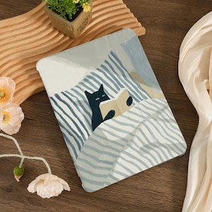 May include: A tablet case featuring an illustration of a black cat reading a book in bed. The design includes blue and white striped bedding and a light blue pillow. The case is on a wooden surface.