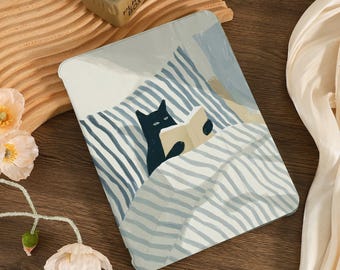 Puede incluir: Una funda para tableta con una ilustración de un gato negro leyendo un libro en la cama. El diseño incluye ropa de cama a rayas azules y blancas y una almohada azul claro. La funda está sobre una superficie de madera.