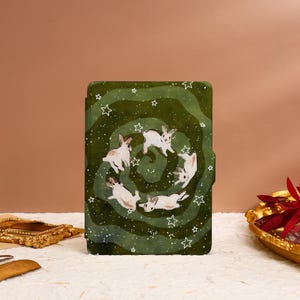 May include: A green notebook cover with a spiral design featuring white rabbits and stars. The cover has a magnetic closure and is set against a neutral background with gold accents.