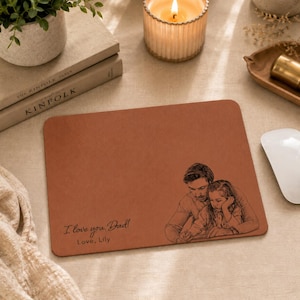 Può includere: Un mousepad rettangolare marrone con uno schizzo di un padre e un bambino. Il testo "I love you, Dad! Love, Lily" è stampato in basso a sinistra. Un mouse per computer bianco è a destra.