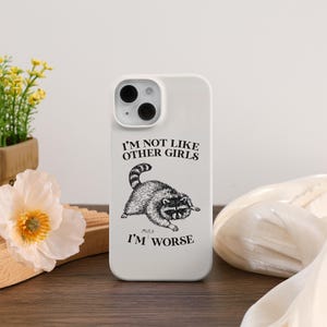 May include: White phone case featuring a raccoon illustration with the text "I'M NOT LIKE OTHER GIRLS MUCH I'M WORSE". The case has a glossy finish and a camera cutout. The phone case is on a wooden surface.