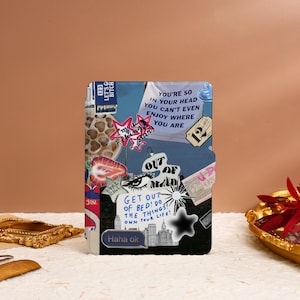 May include: A black notebook with a collage of colorful stickers and text. The stickers include a giraffe print, a strawberry, a star, a cityscape, and a pair of eyes. The text includes "You're so in your head you can't even enjoy where you are", "Get out of bed! Do the things! Own your life!" and "Haha ok".