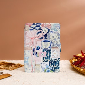 May include: A white notebook with a collage of blue and pink illustrations. The cover features a cup of coffee, a pink bow, and the text "The Little Thing".