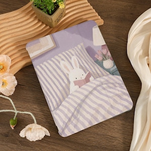May include: A tablet case with a charming illustration of a white rabbit reading in bed. The design features a purple striped duvet, a bedside table with flowers, and a soft colour palette.