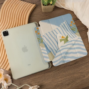 Puede incluir: Un iPad verde claro con una funda transparente, junto a una funda con un pato leyendo un libro en la cama. La funda tiene un patr&oacute;n de rayas azules y blancas, con flores naranjas. El iPad est&aacute; sobre una superficie de madera.