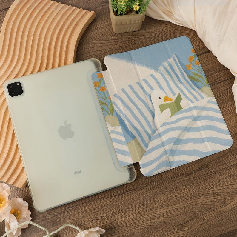 Puede incluir: Un iPad verde claro con una funda transparente, junto a una funda con un pato leyendo un libro en la cama. La funda tiene un patr&oacute;n de rayas azules y blancas, con flores naranjas. El iPad est&aacute; sobre una superficie de madera.