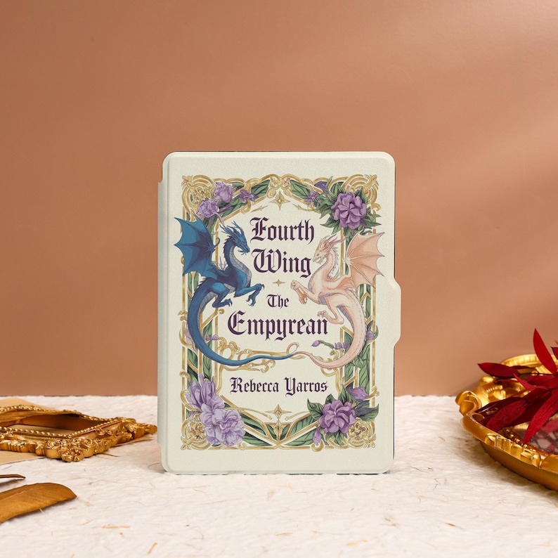 May include: A white book cover with a gold frame and floral design. The title is "Fourth Wing: The Empyrean" by Rebecca Yarros. Two dragons, one blue and one pink, are featured on the cover.