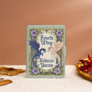 Könnte beinhalten: Ein Buchcover mit grünem Hintergrund und goldenen Akzenten. Der Titel "Fourth Wing" ist in einer dekorativen Schrift geschrieben. Zwei Drachen, einer blau und einer rosa, stehen sich gegenüber. Der Name des Autors, "Rebecca Yarros", steht unter den Drachen.