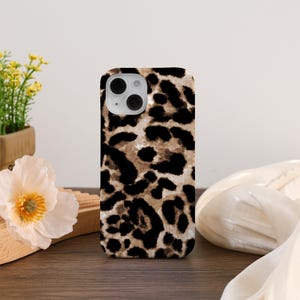 May include: A phone case with a leopard print design. The case features a pattern of black spots on a tan and cream background. The phone case has a camera cutout and is displayed on a wooden surface.
