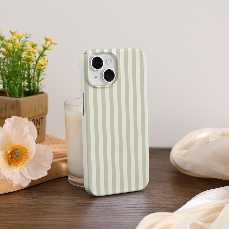 May include: A light green and white striped phone case. The case is on a wooden surface next to a glass of candle and a small potted plant with yellow flowers. A white flower and a piece of fabric are also in the image.