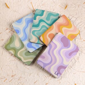 May include: Five rectangular e-reader cases arranged in a fan pattern, each featuring a distinct wavy, multi-coloured striped design. The colours include shades of blue, teal, orange, yellow, green, and purple. One case displays the initials LK.