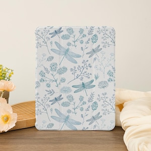 Puede incluir: Una funda para tablet blanca con un patrón de libélula azul y flores. La funda tiene un fondo blanco con ilustraciones azules de libélulas y flores.