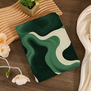 May include: A green and cream-colored notebook with a layered, abstract design. The cover features a pattern of curved lines in varying shades of green and cream. The notebook is rectangular and appears to be closed.