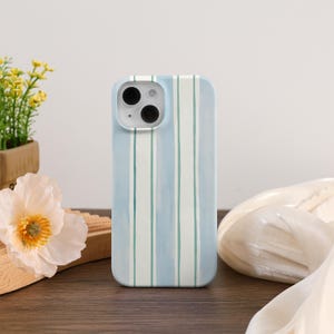 May include: A light blue phone case with vertical stripes in white and teal. The case has a rounded rectangular shape and features a camera cutout. The background includes a wooden surface, flowers, and a white fabric.