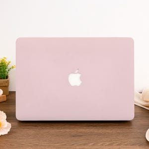 May include: A light pink laptop with the Apple logo in the center. The laptop is closed and sits on a wooden surface. The background is white.