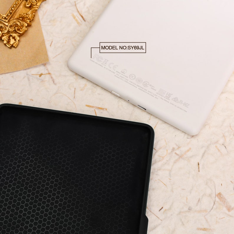 May include: A white e-reader with the model number "SY69JL" printed on the back. The e-reader is lying on a light brown surface. A black case with a honeycomb pattern is next to the e-reader.