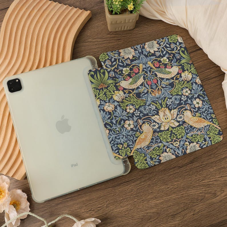 May include: An iPad in a clear case next to a patterned cover. The cover features a dark blue background with a floral and bird design in green, white, and yellow. The iPad is light green.