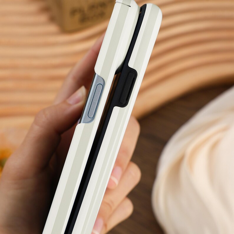 May include: A cream-colored phone case with vertical striped detailing. The case is open, revealing a black interior and a silver-colored button. The phone case is held in a person's hand.